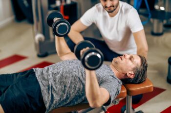 Man in gym with coach doing dumbbell chest press as exercise after prostate cancer diagnosis